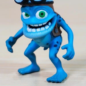 Könnte beinhalten: Blaue Figur einer Cartoon-Froschfigur mit breitem Grinsen und grünen Augen. Der Frosch trägt einen braunen Fliegerhut und eine Weste. Die Figur hat blaue Flecken und befindet sich in einer spielerischen Pose.