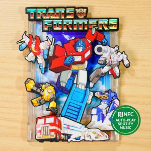May include: A clear plastic case featuring a colorful Transformers design. The case showcases various robot characters in red, blue, yellow, and white. The Transformers logo is at the top. A green circle with the text "NFC AUTO-PLAY SPOTIFY MUSIC" is on the bottom right.