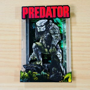 Może przedstawiać: Figurka Predatora w przezroczystym plastikowym etui. Figurka kuca na skalistej podstawie z tłem dżungli. Słowo "PREDATOR" jest w czerwonych blokowych literach u góry.