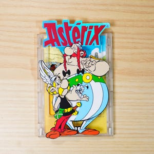 May include: A clear plastic case with a vibrant Asterix and Obelix design. The image shows the characters with the text "Asterix" in red and blue. The background depicts a sunny landscape with a blue sky.