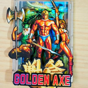 May include: A vibrant Golden Axe video game cartridge featuring three muscular characters with weapons. The title "GOLDEN AXE" is displayed in bold, pixelated letters. The artwork depicts a warrior with an axe, a blonde warrior with a sword, and a woman with a sword.