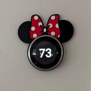 以下が含まれることがあります： 73の数字が表示された黒い画面のスマートサーモスタット。黒いミニーマウスをテーマにしたフレームで囲まれています。フレームには黒いミッキーマウスの耳と、白い水玉模様の赤いリボンが付いています。