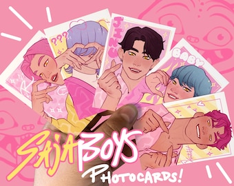 May include: A hand holding a collection of photocard illustrations featuring stylized portraits of young men with various hair colors. The cards are set against a pink background with the text "SAJA BOYS PHOTOCARDS!"