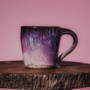 May include: A ceramic mug featuring a reactive glaze with a gradient of deep purple, magenta, and white tones. The mug has a thick, curved handle and a textured, unglazed base. The surface displays a glossy finish with visible color blending.