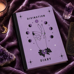 May include: A light purple hardcover diary with the text DIVINATION DIARY printed on the front cover. The cover features a central illustration of a hand surrounded by a circular moon phase cycle, accented with small stars and a floral wrist detail.