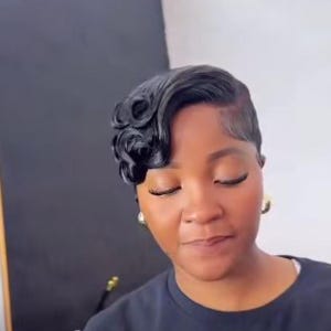 May include: A woman with short, styled black hair. The hair is styled in waves on one side and closely cropped on the other. The woman has closed eyes and is wearing gold earrings.