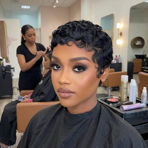 May include: A woman with short, dark, wavy hair and full makeup, including dark eyeshadow and lipstick, is the focus. She is wearing a black salon cape. A stylist is working on another client in the background.