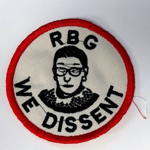 Può includere: Una toppa circolare bianca con un bordo rosso. La toppa presenta un'illustrazione in bianco e nero di Ruth Bader Ginsburg, con il testo "RBG" sopra di lei e "WE DISSENT" sotto.