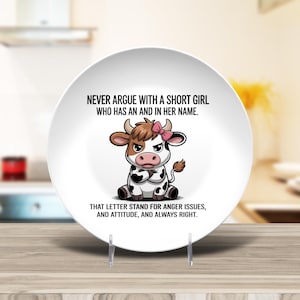 May include: A white ceramic plate with a cartoon cow illustration and text. The cow has a pink bow and an angry expression. The text reads: "NEVER ARGUE WITH A SHORT GIRL WHO HAS AN AND IN HER NAME. THAT LETTER STAND FOR ANGER ISSUES, AND ATTITUDE, AND ALWAYS RIGHT."