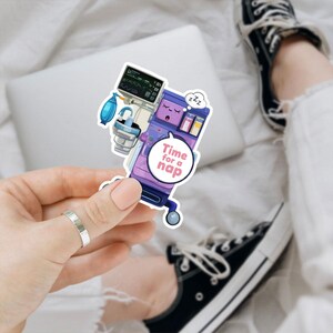 May include: A sticker featuring a cartoon illustration of an anesthesia machine in shades of purple, blue, and white. The sticker has the text "Time for a nap" in a speech bubble. The machine has a sleeping face and a "zzz" symbol.