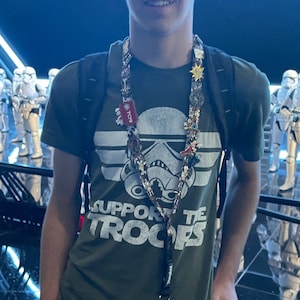 May include: A young person wearing a green t-shirt with the text "Support the Troops" and a white stormtrooper graphic. They are wearing a lanyard with multiple pins and charms. The person is standing in front of a backdrop of white stormtroopers.