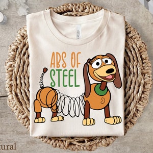 Puede incluir: Una camiseta beige con el texto "Abs of Steel" y una imagen de dibujos animados de un perro marr&oacute;n y naranja con un muelle para la cola. El perro lleva un collar verde.