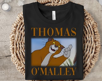 May include: Black t-shirt featuring a cartoon image of Thomas O'Malley, a brown cat with a friendly expression, waving his paw. The name "THOMAS O'MALLEY" is printed in orange above and below the image. The shirt is folded on a woven surface.