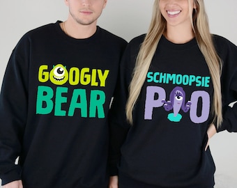 May include: Two black sweatshirts with a graphic print of characters from the movie Monsters Inc. The sweatshirt on the left says "Googly Bear" in green and yellow text with a green monster with one eye. The sweatshirt on the right says "Schmoopsie P.O." in purple and teal text with a purple monster with one eye.