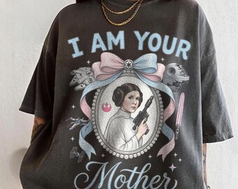 May include: Dark grey t-shirt featuring a portrait of Princess Leia holding a blaster, surrounded by a pink and blue ribbon and the text "I AM YOUR Mother". The shirt also includes Star Wars imagery.