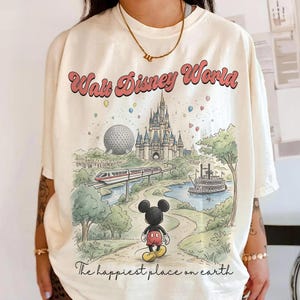 May include: Cream-colored t-shirt featuring a colorful illustration of Walt Disney World. The graphic includes the park's castle, monorail, and Mickey Mouse. Text reads "Walt Disney World" and "The happiest place on earth."