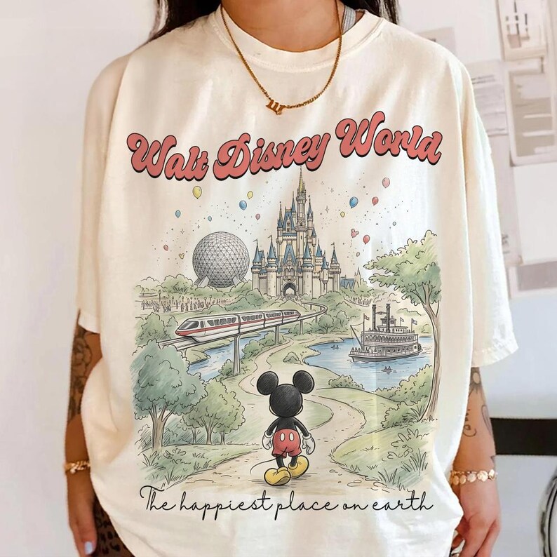 May include: Cream-colored t-shirt featuring a colorful illustration of Walt Disney World. The graphic includes the park's castle, monorail, and Mickey Mouse. Text reads "Walt Disney World" and "The happiest place on earth."