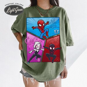 May include: Light green t-shirt featuring a comic book style graphic of Spider-Man and other characters. The graphic is divided into four sections with a blue, red, and pink color scheme. The text "COMFORT COLORS" and "Light Green" is printed on the top left.