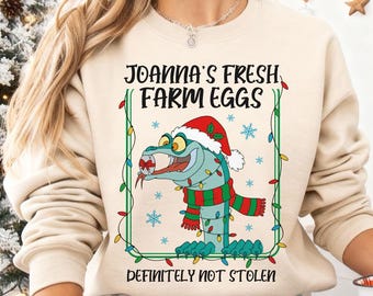 May include: A beige sweatshirt with a Christmas-themed graphic. The graphic features a cartoon snake wearing a Santa hat and a scarf, surrounded by Christmas lights and snowflakes. Text reads "JOANNA'S FRESH FARM EGGS" and "DEFINITELY NOT STOLEN".