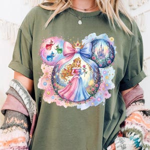 May include: Olive green t-shirt with a vibrant graphic of a princess in a pink and blue gown holding a rose. The design includes a castle, fairies, and a large pink and blue bow, all within a Mickey Mouse ear shape.