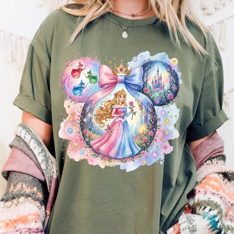 May include: Olive green t-shirt with a vibrant graphic of a princess in a pink and blue gown holding a rose. The design includes a castle, fairies, and a large pink and blue bow, all within a Mickey Mouse ear shape.
