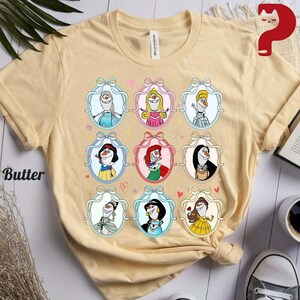 May include: Butter-colored t-shirt featuring a design of cartoon characters in oval frames with bows and hearts. The characters are in various outfits and poses. The shirt is made of a soft material.