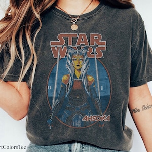 Op de afbeelding: Een donkergrijs T-shirt met een vintage-stijl afbeelding van Ahsoka Tano uit Star Wars. De afbeelding toont Ahsoka met haar lichtzwaarden en de tekst "Star Wars" en "Ahsoka".