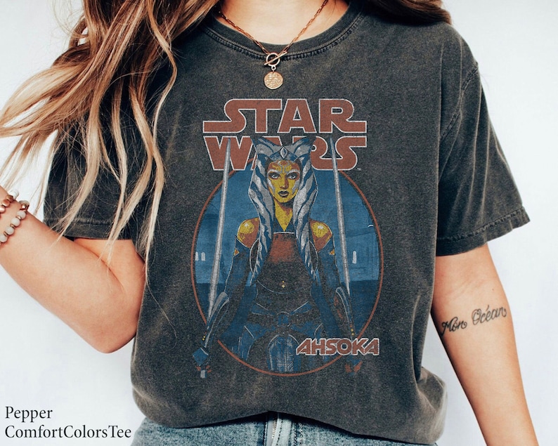 Op de afbeelding: Een donkergrijs T-shirt met een vintage-stijl afbeelding van Ahsoka Tano uit Star Wars. De afbeelding toont Ahsoka met haar lichtzwaarden en de tekst "Star Wars" en "Ahsoka".