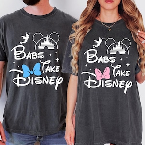 May include: Two dark gray t-shirts with the text "Babs Take Disney" in white script. One shirt has a blue bow, the other a pink bow. The design includes a castle, Mickey Mouse ears, a fairy, and stars.
