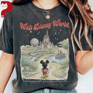 May include: Dark gray t-shirt featuring a colorful illustration of Walt Disney World. The design includes the park's castle, a monorail, and Mickey Mouse. The text "Walt Disney World" and "The happiest place on earth" are also visible.