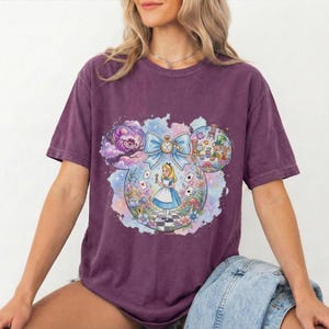 May include: A purple t-shirt featuring a colorful graphic of Alice in Wonderland in a Mickey Mouse-shaped design. The design includes the Cheshire Cat, Alice, and other iconic elements from the story.