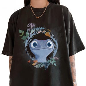 Puede incluir: Camiseta negra con un dise&ntilde;o central de una criatura azul parecida a una salamandra, con grandes ojos azules, rodeada por una corona de hojas y flores. El dise&ntilde;o es un elemento visual llamativo.