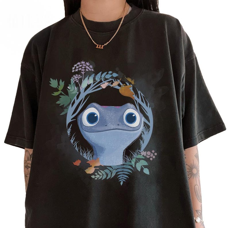 Puede incluir: Camiseta negra con un dise&ntilde;o central de una criatura azul parecida a una salamandra, con grandes ojos azules, rodeada por una corona de hojas y flores. El dise&ntilde;o es un elemento visual llamativo.