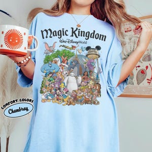 May include: Light blue t-shirt featuring a colorful graphic of Disney characters with the text "Magic Kingdom Walt Disney World." The shirt also has the text "Comfort Colors Chambray" on the bottom left. A red and white mug with a sun design is held in the image.