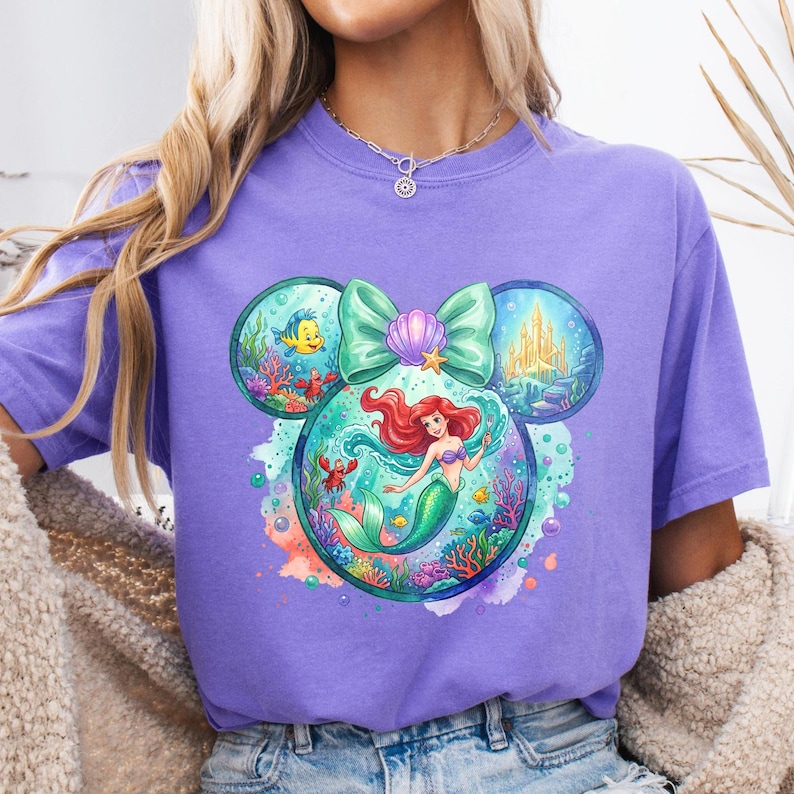 May include: A lilac t-shirt with a vibrant graphic of Ariel from The Little Mermaid. The design incorporates a bow, a seashell, and images of Flounder, a crab, and a castle. The graphic is set within a Mickey Mouse ear shape.