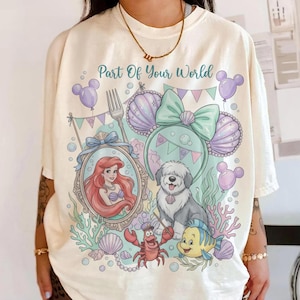 Może przedstawiać: Kremowa koszulka z kolorową grafiką z napisem "Part Of Your World". Projekt zawiera Ariel, psa, kraba i rybę, a także elementy muszli i uszu Myszki Miki. Koszulka ma luźny kr&oacute;j.
