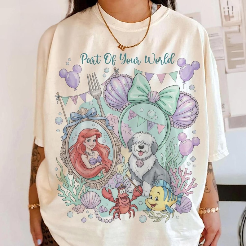 Może przedstawiać: Kremowa koszulka z kolorową grafiką z napisem "Part Of Your World". Projekt zawiera Ariel, psa, kraba i rybę, a także elementy muszli i uszu Myszki Miki. Koszulka ma luźny kr&oacute;j.