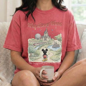May include: Red t-shirt featuring a graphic of Mickey Mouse in front of a Disney World scene, including a castle, monorail, and the text "Walt Disney World" and "The happiest place on earth". The person is holding a beige mug.