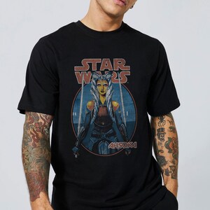 Op de afbeelding: Zwart T-shirt met een vintage-stijl afbeelding van Ahsoka Tano uit Star Wars. De afbeelding toont Ahsoka met twee lichtzwaarden en de tekst "STAR WARS" en "AHSOKA".