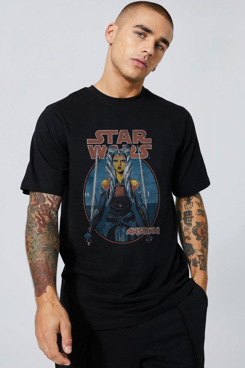 Op de afbeelding: Zwart T-shirt met een vintage-stijl afbeelding van Ahsoka Tano uit Star Wars. De afbeelding toont Ahsoka met twee lichtzwaarden en de tekst "STAR WARS" en "AHSOKA".
