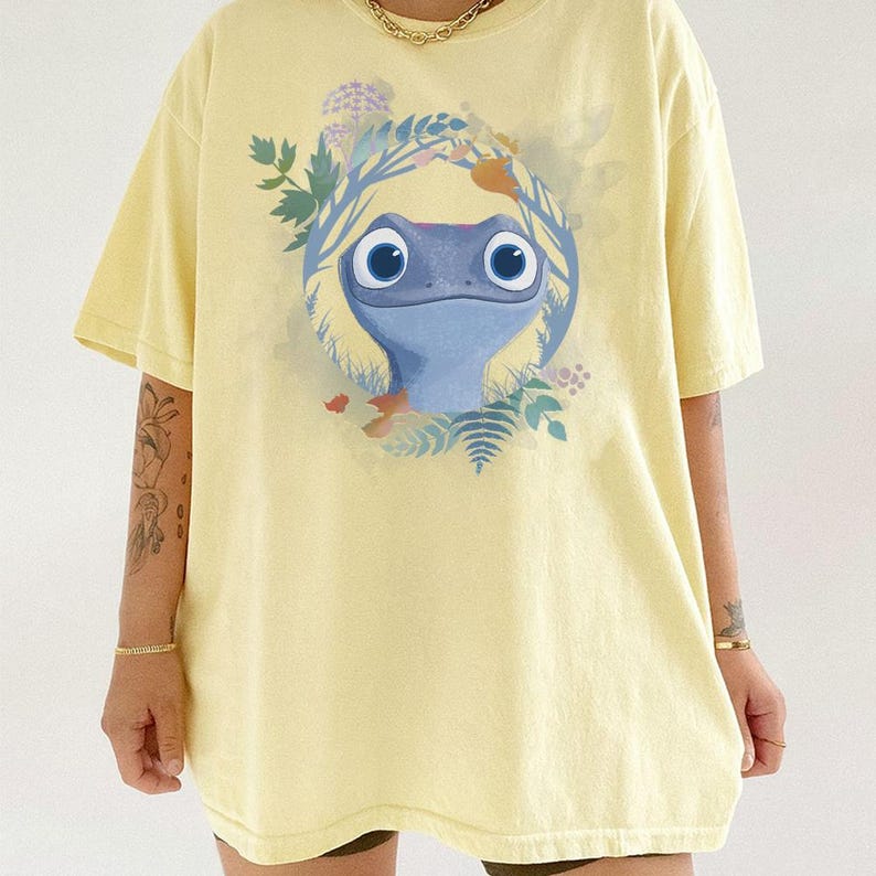 Puede incluir: Camiseta amarilla clara con un dise&ntilde;o de lagarto de dibujos animados azul, rodeado por una corona floral. La camiseta tiene mangas cortas y es de color liso.
