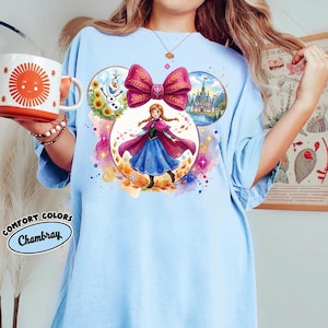 May include: Pale blue t-shirt with a vibrant graphic of a princess, a large pink bow, a castle, Olaf, and sunflowers. The shirt also has the words "Comfort Colors Chambray" printed on it.