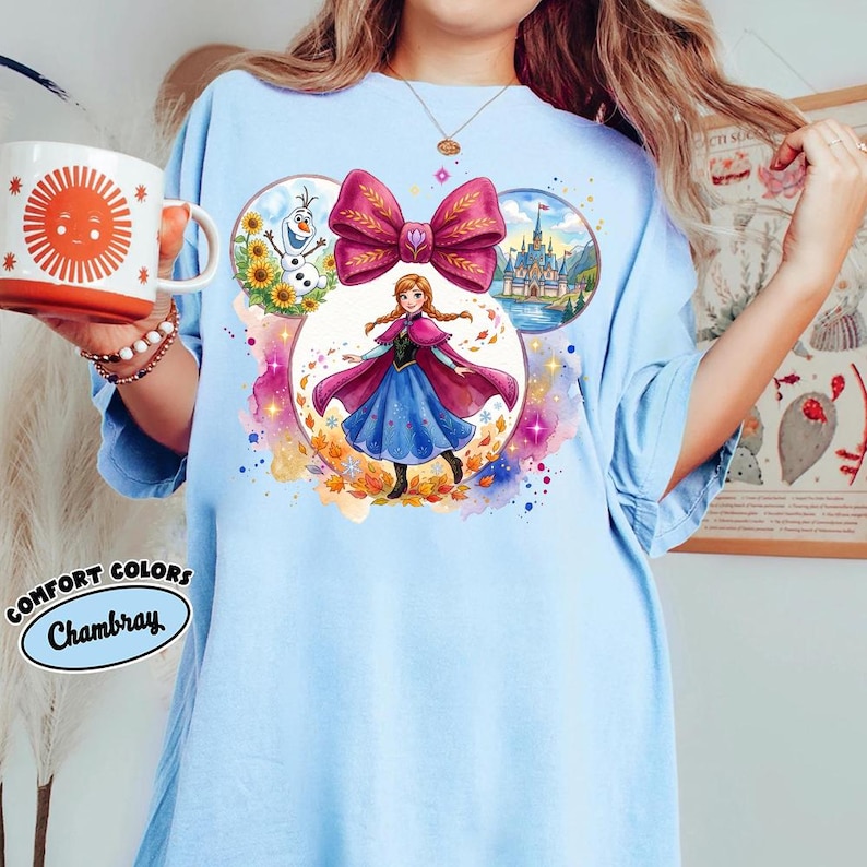 May include: Pale blue t-shirt with a vibrant graphic of a princess, a large pink bow, a castle, Olaf, and sunflowers. The shirt also has the words "Comfort Colors Chambray" printed on it.