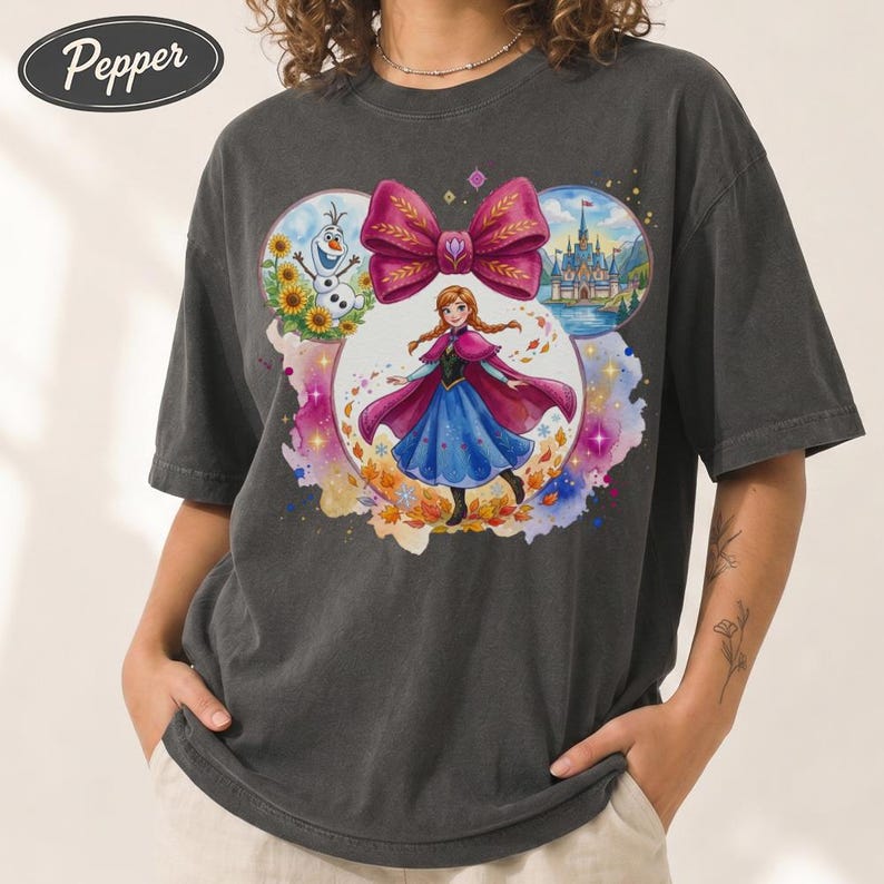 Puede incluir: Camiseta gris oscuro con un gr&aacute;fico colorido de Anna de Frozen, un gran lazo rosa e im&aacute;genes de Olaf y un castillo. La camiseta tiene un corte holgado y mangas cortas. La palabra "Pepper" est&aacute; impresa en la parte superior izquierda.