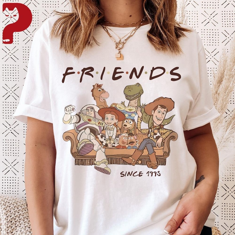 May include: White t-shirt featuring the word "FRIENDS" in brown letters above a graphic of Toy Story characters sitting on a couch. The phrase "SINCE 1995" is printed below the graphic. The shirt has short sleeves.
