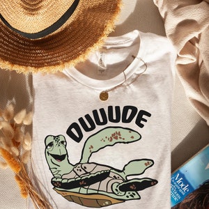 以下が含まれることがあります： 白いTシャツに、緑色のウミガメの漫画と黒い文字で「DUUUDE」と書かれています。ウミガメは笑顔で、茶色の甲羅をしています。