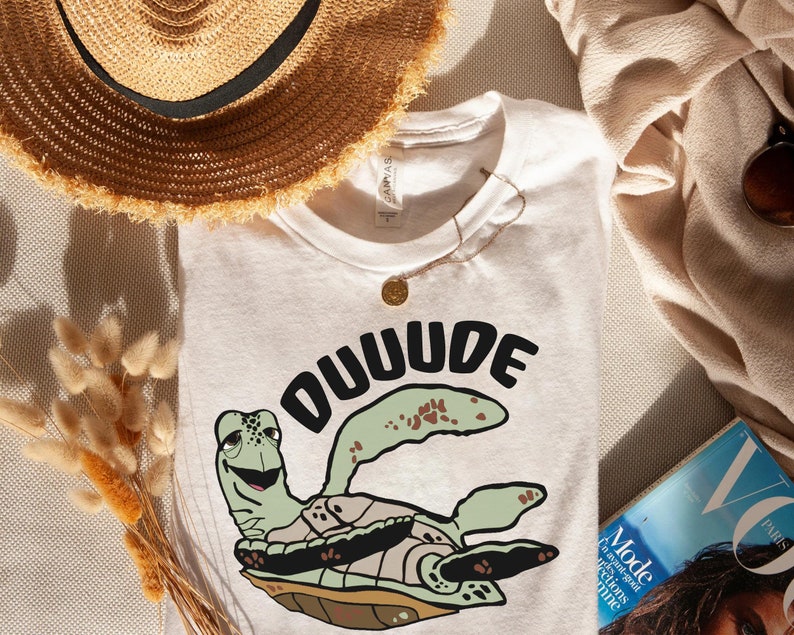 以下が含まれることがあります： 白いTシャツに、緑色のウミガメの漫画と黒い文字で「DUUUDE」と書かれています。ウミガメは笑顔で、茶色の甲羅をしています。