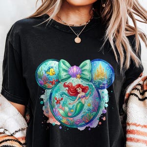 May include: Black t-shirt with a colourful graphic of Ariel the mermaid within a Mickey Mouse ear design. The design includes a teal bow, a seashell, and underwater scenes with fish and a castle.