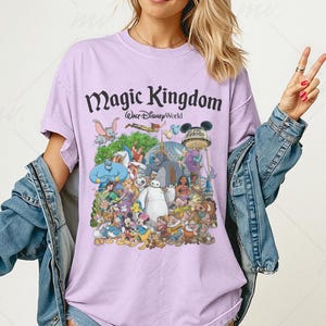 May include: A lilac-colored t-shirt featuring a colorful graphic of various Disney characters, including Mickey Mouse, Dumbo, and Baymax. The text "Magic Kingdom Walt Disney World" is printed across the top in a stylized font.