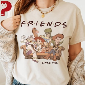 May include: Beige t-shirt featuring the word "FRIENDS" in brown letters above a graphic of Toy Story characters sitting on a couch. The text "SINCE 1995" is printed below the couch in brown letters.